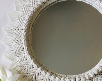 May include: A round, white macrame mirror with a sunburst design. The mirror's frame is intricately woven with a geometric pattern and fringed edges. A few white flowers are visible in the lower left corner, adding a touch of elegance.