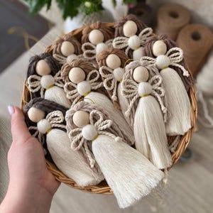 Pode incluir: Uma coleção de enfeites de anjos feitos à mão em uma cesta de vime. Cada anjo apresenta uma cabeça de contas de madeira, cabelo de fio e um laço de fio com asas esvoaçantes. Os anjos têm cores de cabelo variadas e são acentuados com fio dourado.