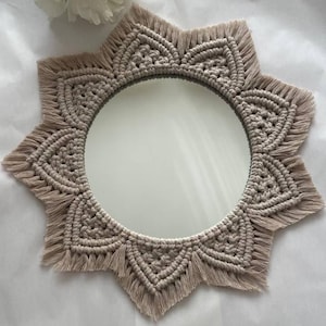 May include: A round mirror with a beige macrame sunburst design. The mirror has a circular reflective surface surrounded by a detailed, woven pattern. The macrame frame has a fringed edge, adding texture and visual interest to the decorative wall hanging.