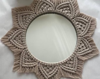 May include: A round mirror with a beige macrame sunburst design. The mirror has a circular reflective surface surrounded by a detailed, woven pattern. The macrame frame has a fringed edge, adding texture and visual interest to the decorative wall hanging.