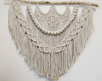 Puede incluir: Colgante de pared de macramé color crema. La pieza hecha a mano presenta intrincados patrones de nudos y un borde con flecos. Está suspendida de un pasador de madera natural, lo que añade un toque rústico a la decoración. El diseño es simétrico y geométrico.