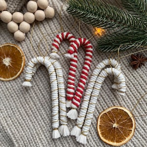 Könnte beinhalten: Nahaufnahme von handgefertigten Weihnachtsschmuck. Drei weiße Zuckerstangen-Ornamente mit goldenen Akzenten und Quasten und eine rot-weiß gestreifte Zuckerstange. Die Ornamente liegen auf einer strukturierten, hellgrauen Oberfläche mit getrockneten Orangenscheiben und Grünzeug.