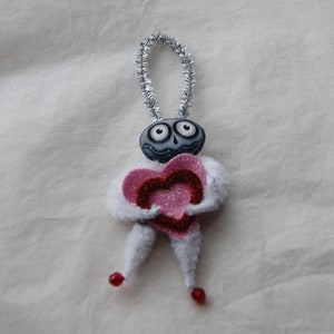 May include: A handmade Valentine's Day ornament featuring a whimsical figure with a gray face, large eyes, and a silver pipe cleaner loop. The figure holds a pink glitter heart and has white fuzzy arms and legs, with red bead feet.