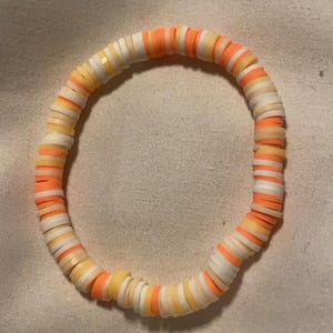 Peut inclure: Un bracelet color&eacute; compos&eacute; de petites perles rondes dans des tons de blanc, jaune et orange. Les perles sont dispos&eacute;es selon un motif r&eacute;p&eacute;titif.