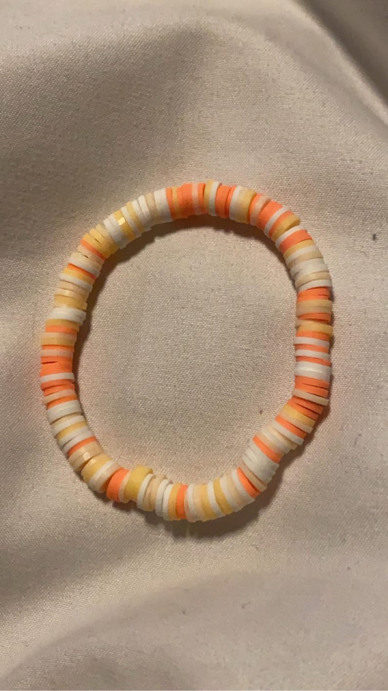 Peut inclure: Un bracelet color&eacute; compos&eacute; de petites perles rondes dans des tons de blanc, jaune et orange. Les perles sont dispos&eacute;es selon un motif r&eacute;p&eacute;titif.
