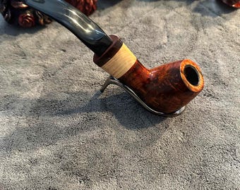 May include: A handcrafted tobacco pipe with a dark, curved stem and a reddish-brown bowl. The bowl has a light-colored band and rests on a metal stand. Several carved wooden heads are in the background.