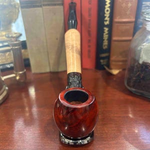 May include: A close-up of a red and black wooden pipe with a bowl and a stem. The pipe is sitting on a wooden surface.