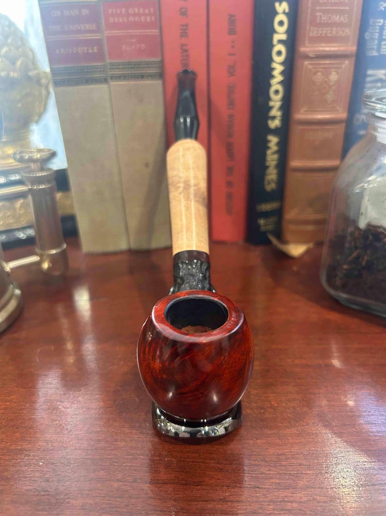 May include: A close-up of a red and black wooden pipe with a bowl and a stem. The pipe is sitting on a wooden surface.