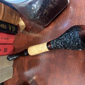 May include: A black and brown wooden smoking pipe with a textured bowl. The pipe has a black stem and a red accent.