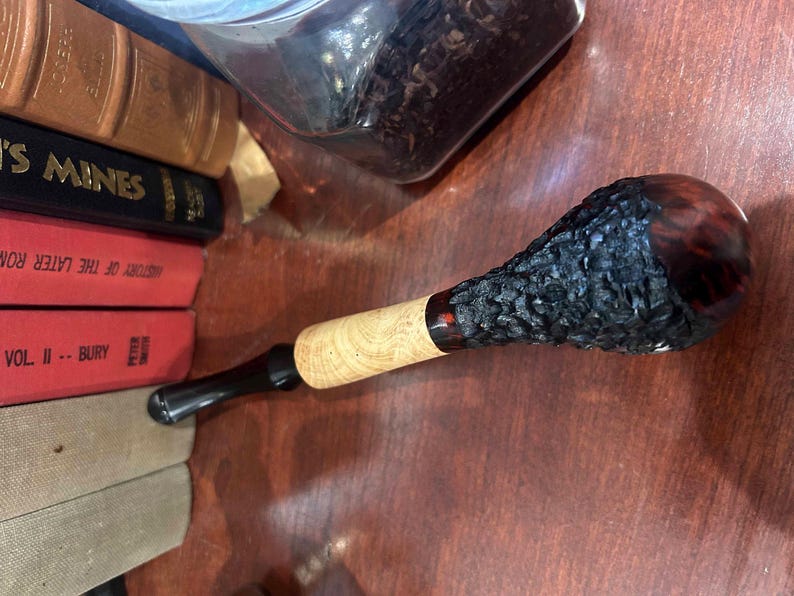 May include: A black and brown wooden smoking pipe with a textured bowl. The pipe has a black stem and a red accent.