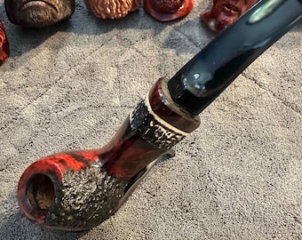 May include: A handcrafted tobacco pipe with a red and black mottled finish. The pipe features a black mouthpiece and a textured bowl. Several carved wooden busts of faces are in the background.