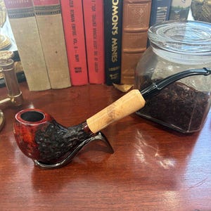 May include: A close-up of a wooden smoking pipe with a red and black bowl and a natural wood stem. The pipe is resting on a wooden surface next to a glass jar filled with dark brown tobacco.