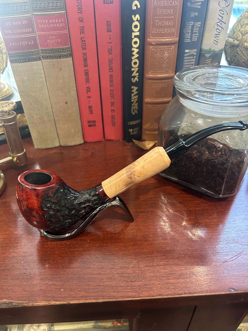 May include: A close-up of a wooden smoking pipe with a red and black bowl and a natural wood stem. The pipe is resting on a wooden surface next to a glass jar filled with dark brown tobacco.