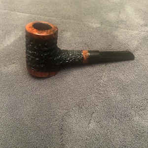 May include: A tobacco pipe with a dark, textured bowl and a black stem. The bowl has a reddish-brown rim and a textured, dark surface. The stem is black and angled. The pipe is designed for smoking tobacco.