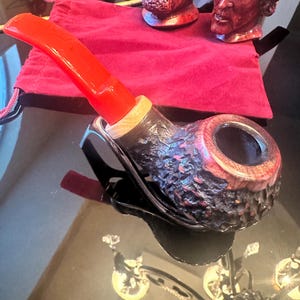 May include: A handcrafted tobacco pipe with a red stem and a dark, textured bowl. The pipe rests on a reflective surface, with two carved wooden busts and a red pouch in the background. The pipe's bowl has a unique, carved design.
