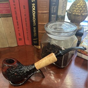 May include: A black and red wooden pipe with a light brown stem. The pipe is sitting on a wooden table next to a glass jar filled with dark brown tobacco.