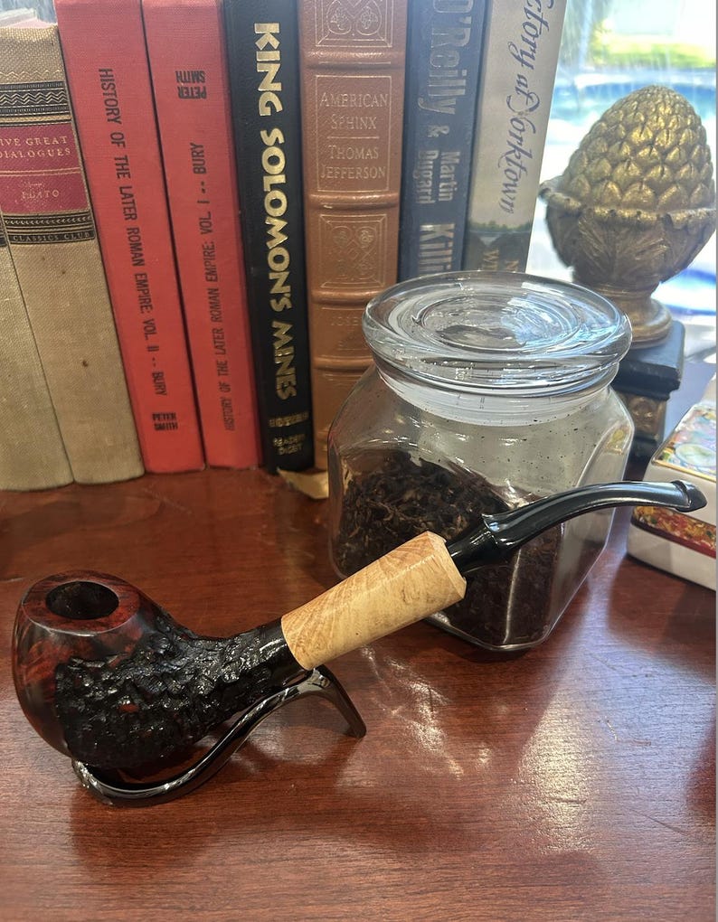 May include: A black and red wooden pipe with a light brown stem. The pipe is sitting on a wooden table next to a glass jar filled with dark brown tobacco.