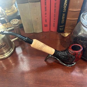 May include: A close-up of a dark brown and red smoking pipe with a black stem and a wooden bowl. The pipe is resting on a wooden surface with a gold metal object and a glass jar in the background.