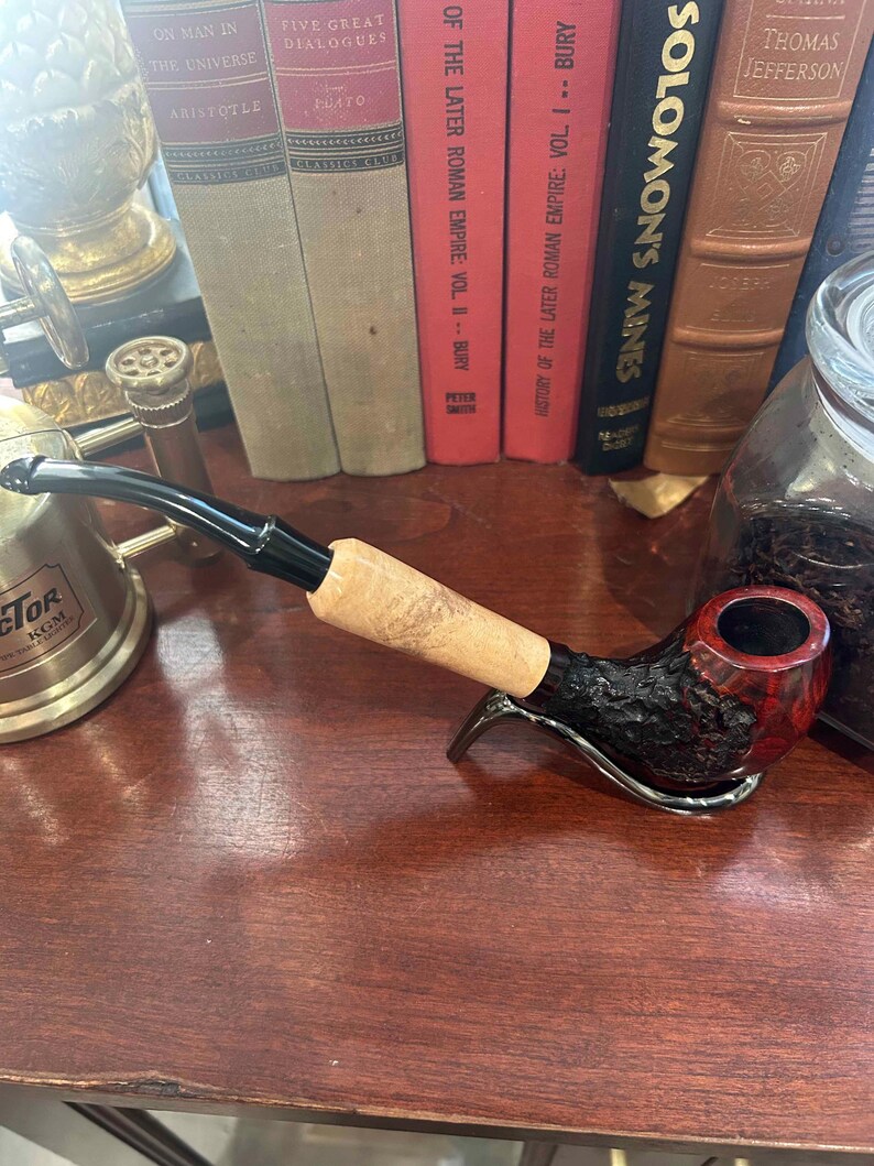 May include: A close-up of a dark brown and red smoking pipe with a black stem and a wooden bowl. The pipe is resting on a wooden surface with a gold metal object and a glass jar in the background.