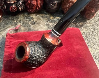May include: A handcrafted tobacco pipe with a black stem and a textured, dark-stained bowl. The bowl is set on a red velvet pouch. Several carved pipes with unique designs are visible in the background.