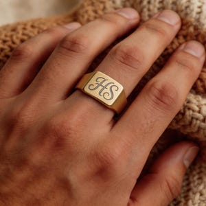 May include: A gold-toned signet ring featuring a flat, rectangular face engraved with the cursive letters HS. The ring has a smooth, polished finish and a solid band design, showcasing a classic style with a personalized monogram detail.