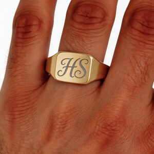 May include: A gold-toned signet ring featuring a flat, rectangular face with rounded corners. The surface of the ring is engraved with the stylized, cursive letters HS in a dark, contrasting tone, centered on the polished metal face.