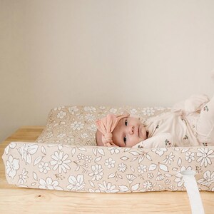 Peut inclure: Un b&eacute;b&eacute; portant un bandeau p&ecirc;che et un ensemble floral de couleur claire est allong&eacute; sur un matelas &agrave; langer. Le matelas &agrave; langer et sa housse ont un fond beige avec des motifs floraux blancs. Le b&eacute;b&eacute; est sur une table &agrave; langer en bois.