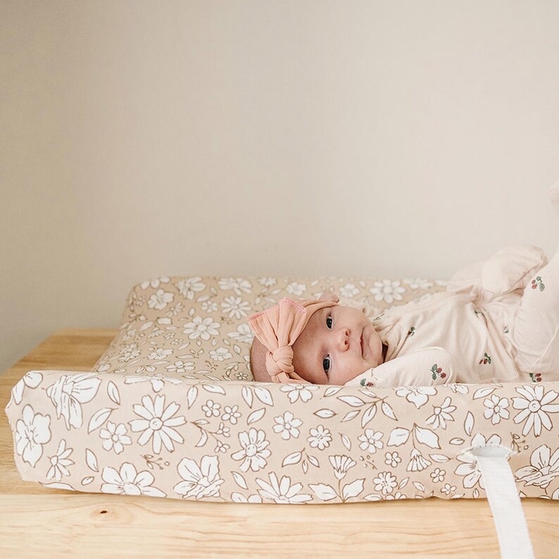 Peut inclure: Un b&eacute;b&eacute; portant un bandeau p&ecirc;che et un ensemble floral de couleur claire est allong&eacute; sur un matelas &agrave; langer. Le matelas &agrave; langer et sa housse ont un fond beige avec des motifs floraux blancs. Le b&eacute;b&eacute; est sur une table &agrave; langer en bois.