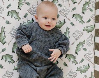 May include: A baby wearing a grey sweater and trousers is lying on a white cot sheet with a green and white fish and fishing net pattern.