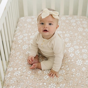 Peut inclure: Un b&eacute;b&eacute; v&ecirc;tu d'un ensemble cr&egrave;me &agrave; manches longues et d'un bandeau assorti avec un n&oelig;ud, assis dans un lit b&eacute;b&eacute; blanc. Le matelas du lit b&eacute;b&eacute; est orn&eacute; d'un motif floral blanc et beige.