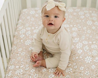 May include: A baby in a cream ribbed onesie and bow headband sits in a white cot. The cot mattress features a floral pattern in white and tan. The baby's outfit has a button-up neckline.