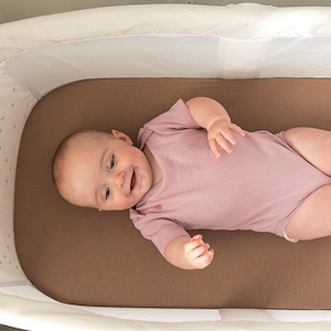 Peut inclure: Un b&eacute;b&eacute; en barboteuse rose est allong&eacute; dans un berceau blanc et marron. Le b&eacute;b&eacute; sourit et regarde vers la cam&eacute;ra. Le berceau a un c&ocirc;t&eacute; en filet et un int&eacute;rieur marron.