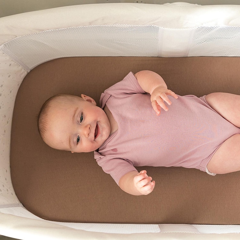 Peut inclure: Un b&eacute;b&eacute; en barboteuse rose est allong&eacute; dans un berceau blanc et marron. Le b&eacute;b&eacute; sourit et regarde vers la cam&eacute;ra. Le berceau a un c&ocirc;t&eacute; en filet et un int&eacute;rieur marron.