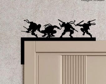 May include: Black metal wall art depicting the Teenage Mutant Ninja Turtles in action. The silhouettes of the four turtles are shown above a door, each holding their distinctive weapons. The artwork is a decorative piece.