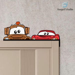 May include: Two cartoon car decorations, one brown and one red, are fixed to the top of a door. The brown car has a truck-like design, while the red car has a sleek, sports car appearance. The decorations are against a grey wall.