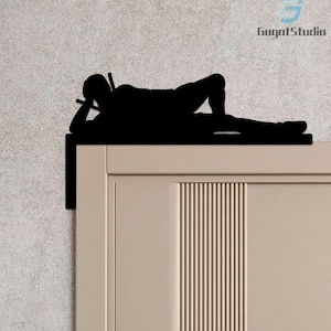 Op de afbeelding: Zwart silhouet van een persoon die op een deurkozijn ligt, met een keu in de hand. De deur is lichtbeige met verticale groeven. De tekst "GogotStudio" staat in de rechterbovenhoek. Een deurdecoratie.