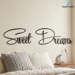May include: Black metal wall art spelling "Sweet Dreams" in a cursive font. The artwork is mounted on a light-coloured wall above a bed with pillows and a decorative shelf.
