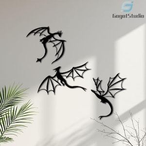 May include: Three black dragon wall art pieces with outstretched wings are mounted on a white wall. The dragons have detailed wing designs and curved tails. The image includes the text "GogotStudio" in the upper right corner. Decorative wall art.