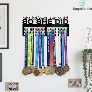 Pode incluir: Expositor de medalhas de metal preto com o texto "She believed she could, so she did". O expositor contém várias medalhas com fitas coloridas. As medalhas são de cor ouro e prata.