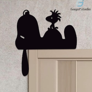 May include: Black silhouette of Snoopy and Woodstock perched on a door frame. The design features Snoopy's iconic form with Woodstock standing on his back. The artwork is mounted on a neutral-colored door, creating a striking contrast.