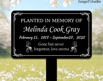 May include: A black memorial plaque with white text, including the name "Melinda Cook Gray" and dates. The plaque reads "Planted in Memory of" and "Gone but never forgotten, love eterna." Decorative floral accents frame the text.