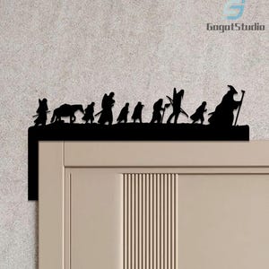 May include: Black metal silhouette of a procession of figures, including a horse, walking across a horizontal bar. The figures are cut out and mounted above a door frame. The text "GogatStudio" is in the upper right corner.