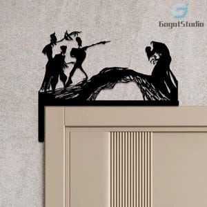 May include: Black metal door topper depicting a silhouette of a dramatic scene with figures on a bridge. The design includes the text "GogotStudio" in the upper right corner. The topper is mounted on a light-coloured door.