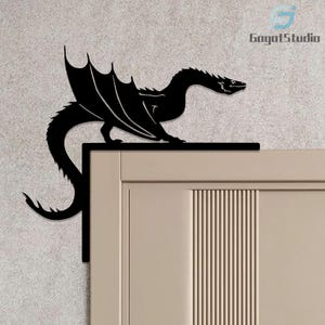 Op de afbeelding: Zwarte metalen drakenwanddecoratie. De draak zit op een hoek, met uitgespreide vleugels en een lange staart. De draak is een silhouet, met een gedetailleerd ontwerp. De wanddecoratie is bevestigd op een lichtbeige deur.
