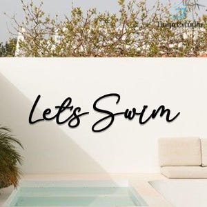 Peut inclure: Décoration murale noire en écriture cursive avec l'inscription "Lets Swim" sur un mur blanc. L'art mural est devant une piscine et un canapé beige. Le haut du mur est orné de verdure.