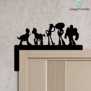 May include: Black metal silhouette featuring characters from a popular animated film. The design includes a dinosaur, a horse, a cowboy, a space ranger, and a speech bubble. The silhouette is mounted on a door frame, adding a decorative touch.