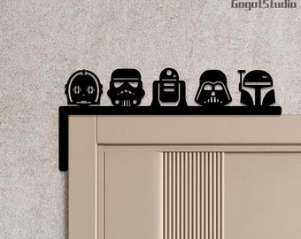 May include: Black metal door topper featuring silhouettes of Star Wars characters: C-3PO, a Stormtrooper, R2-D2, Darth Vader, and Boba Fett. The topper is mounted on a beige door, adding a touch of sci-fi to any room.