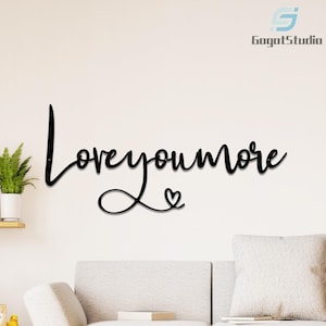 May include: Black metal wall art spelling out "Love you more" in a cursive font, with a small heart symbol. The artwork is mounted on a white wall above a grey sofa. The logo "GogotStudio" is in the upper right corner.