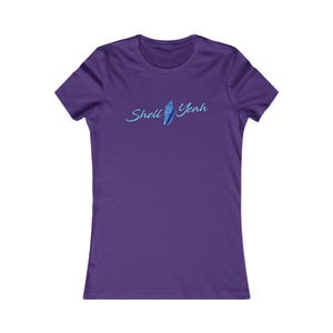 May include: A purple t-shirt featuring the words "Shell Yeah" in light blue script, accompanied by a seashell design. The top has a round neck and short sleeves.