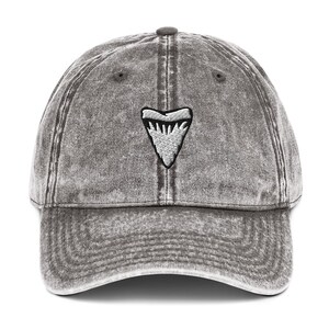 May include: A grey, distressed baseball cap featuring a white shark tooth emblem. The cap has a curved peak and a fabric strap. The hat is made of a soft, worn material, with a vintage look.